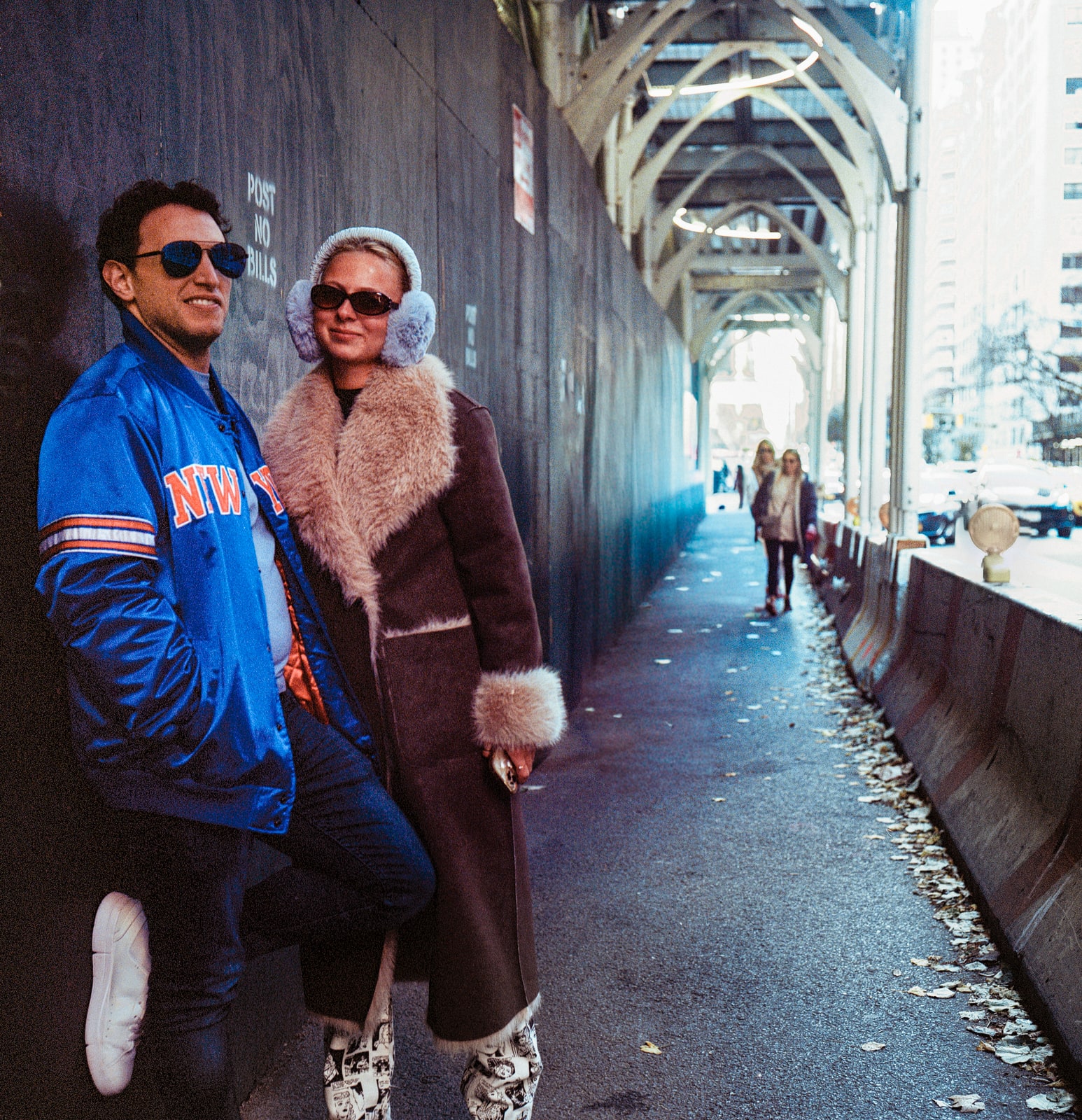 Two people in sunglasses pose beneath steel bridge beams, one in a blue jacket and the other in a brown coat and earmuffs.