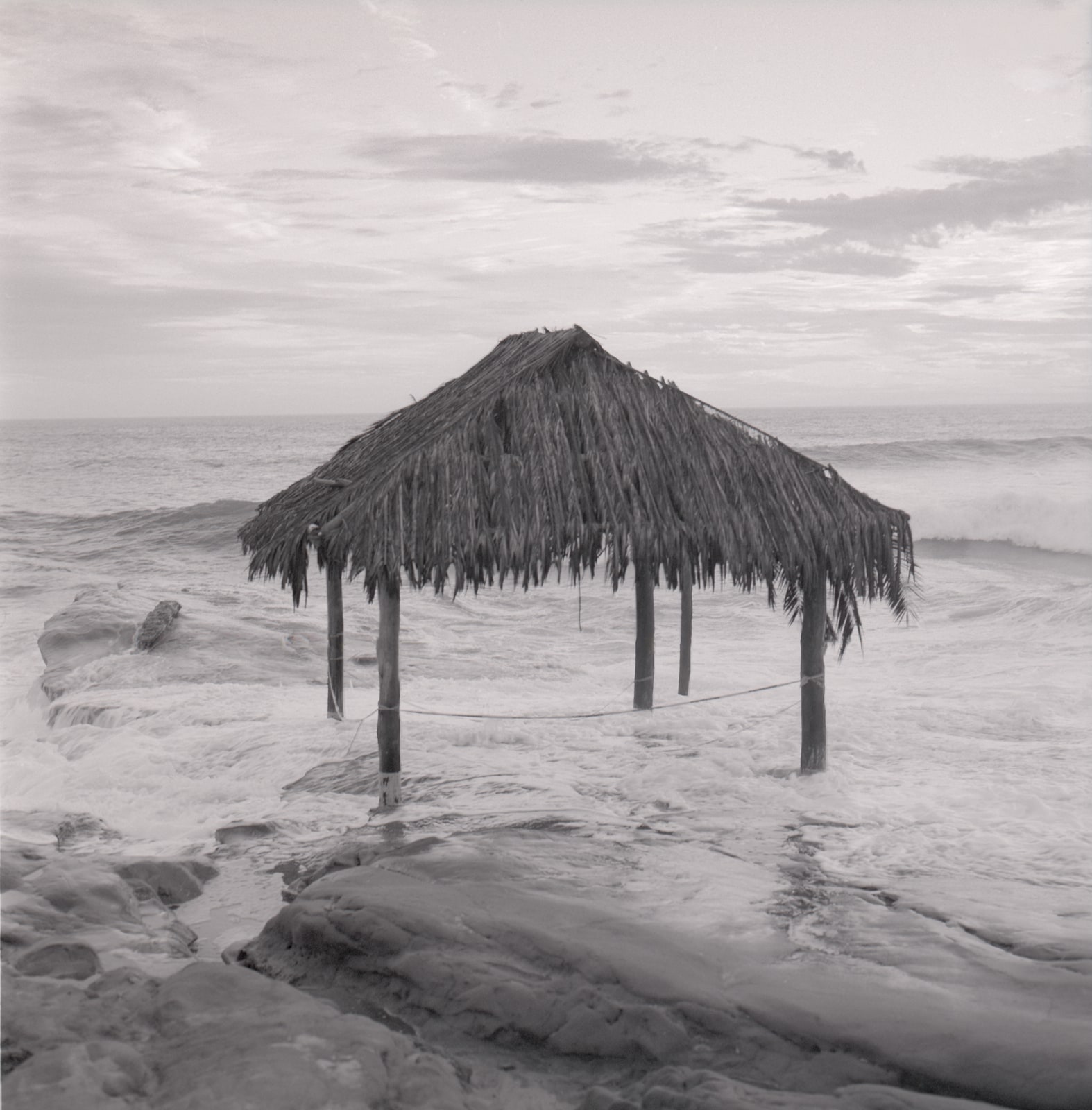 Black-and-white photograph of a thatched-roof shelter standing among coastal rocks, surrounded by breaking ocean waves beneath a cloudy sky.