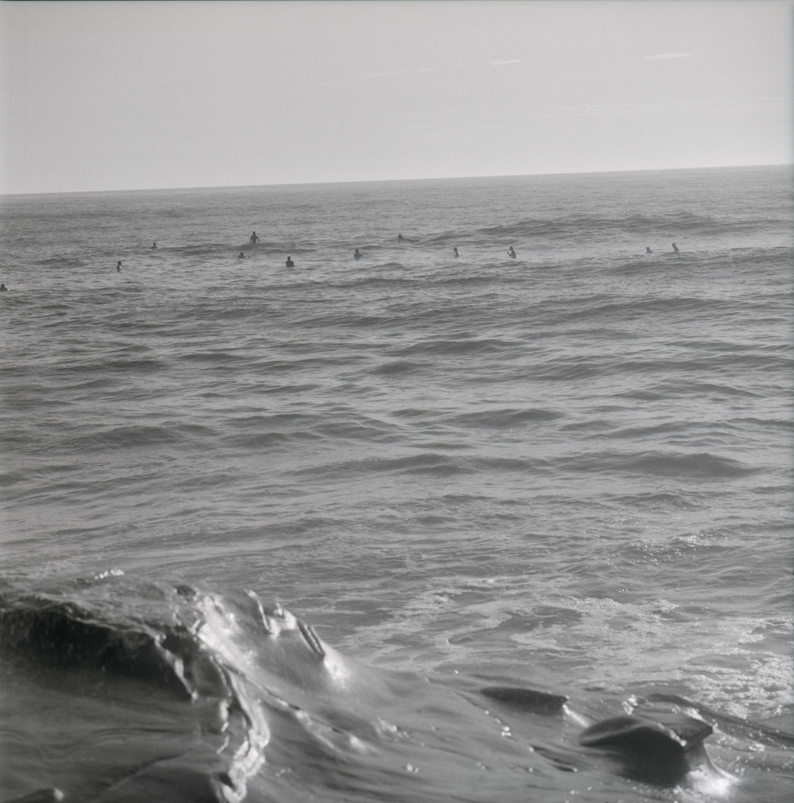 Black-and-white photograph of surfers waiting beyond breaking waves, with rolling ocean swells in the foreground beneath a pale sky.